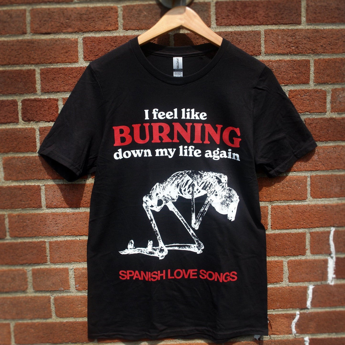 image of a black tee shirt hanging in front of a brick background. front of tee has full body chest print that says i feel like burning down my life again at the top, with a white skeleton kneeling below in the center of the shirt and spanish love songs along the bottom