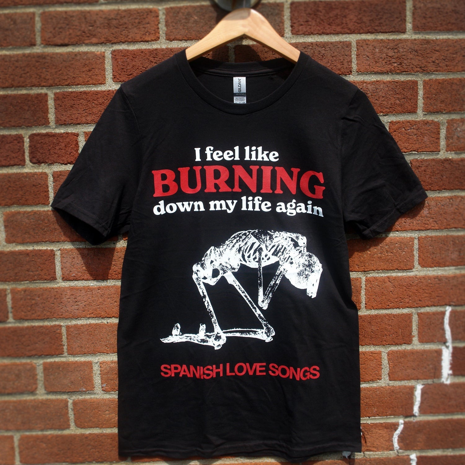 image of a black tee shirt hanging in front of a brick background. front of tee has full body chest print that says i feel like burning down my life again at the top, with a white skeleton kneeling below in the center of the shirt and spanish love songs along the bottom