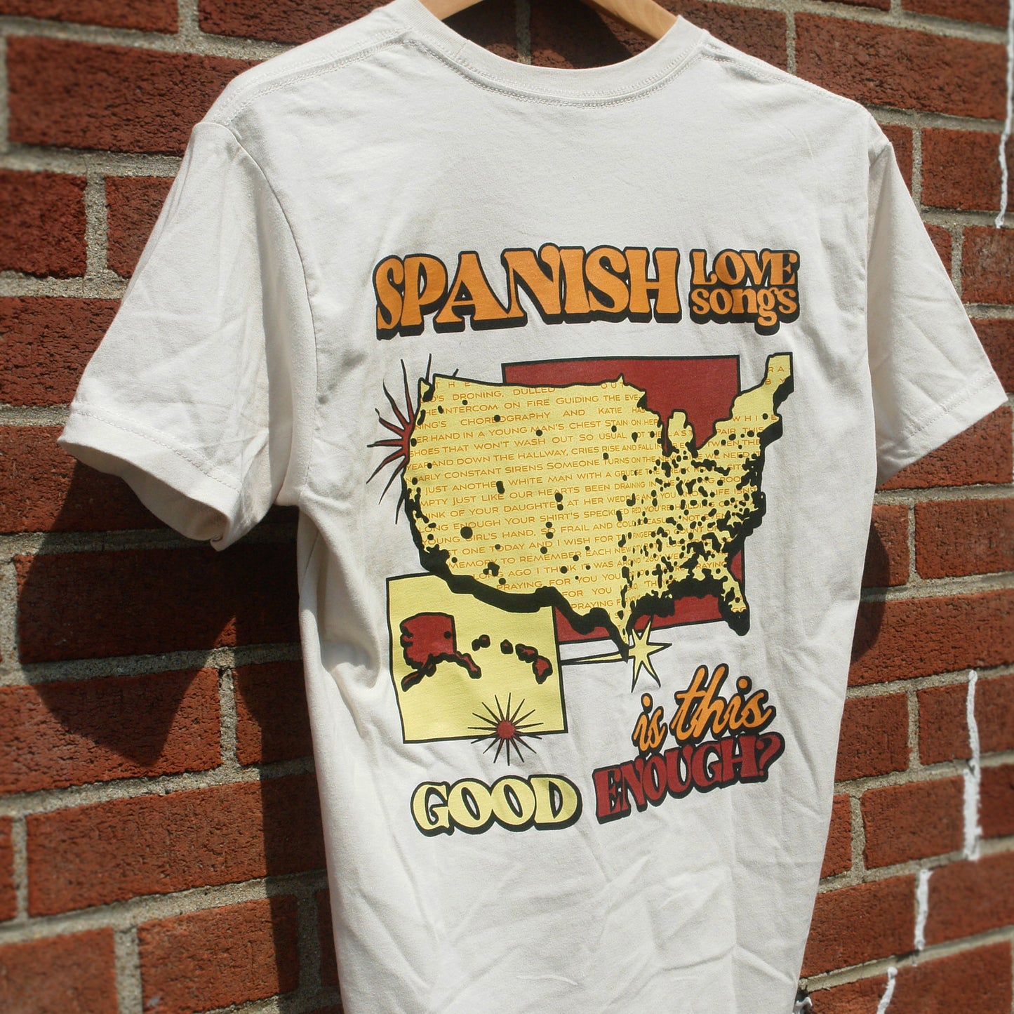 image of the back of a sand colored tee shirt hanging in front of a brick background. the tee has a full back print that has on top in orange in spanish love songs. below that in the center is a map of the united states in yellow and below that in yellow, orange and red says is this good enough?
