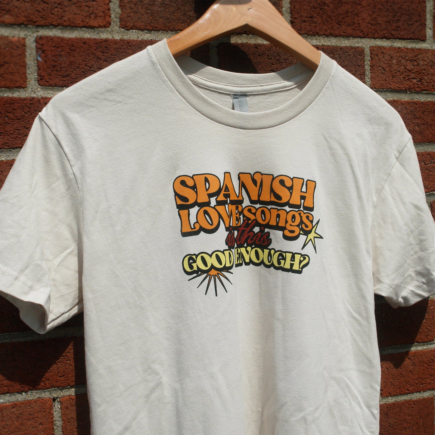 image of the front of a sand colored tee shirt hanging in front of a brick background. the tee has a small center chest print in orange, red and yellow ink that says spanish love songs is this good enough?