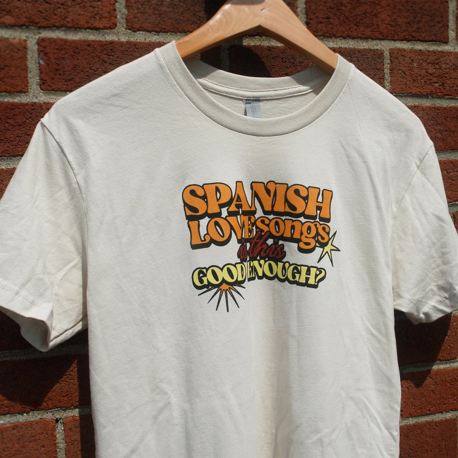 image of the front of a sand colored tee shirt hanging in front of a brick background. the tee has a small center chest print in orange, red and yellow ink that says spanish love songs is this good enough?
