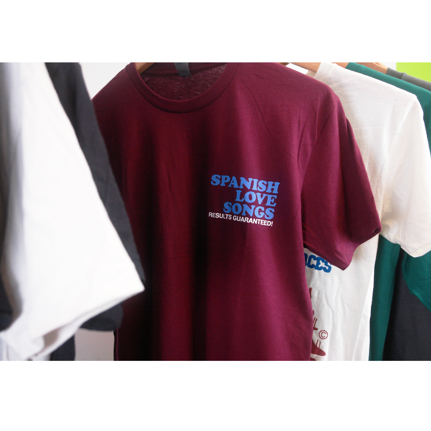 image of the front of a maroon tee shirt on a white background. front of tee has a small chest print on the right. stacked in blue says spanish love songs and below in white says results guaranteed!
