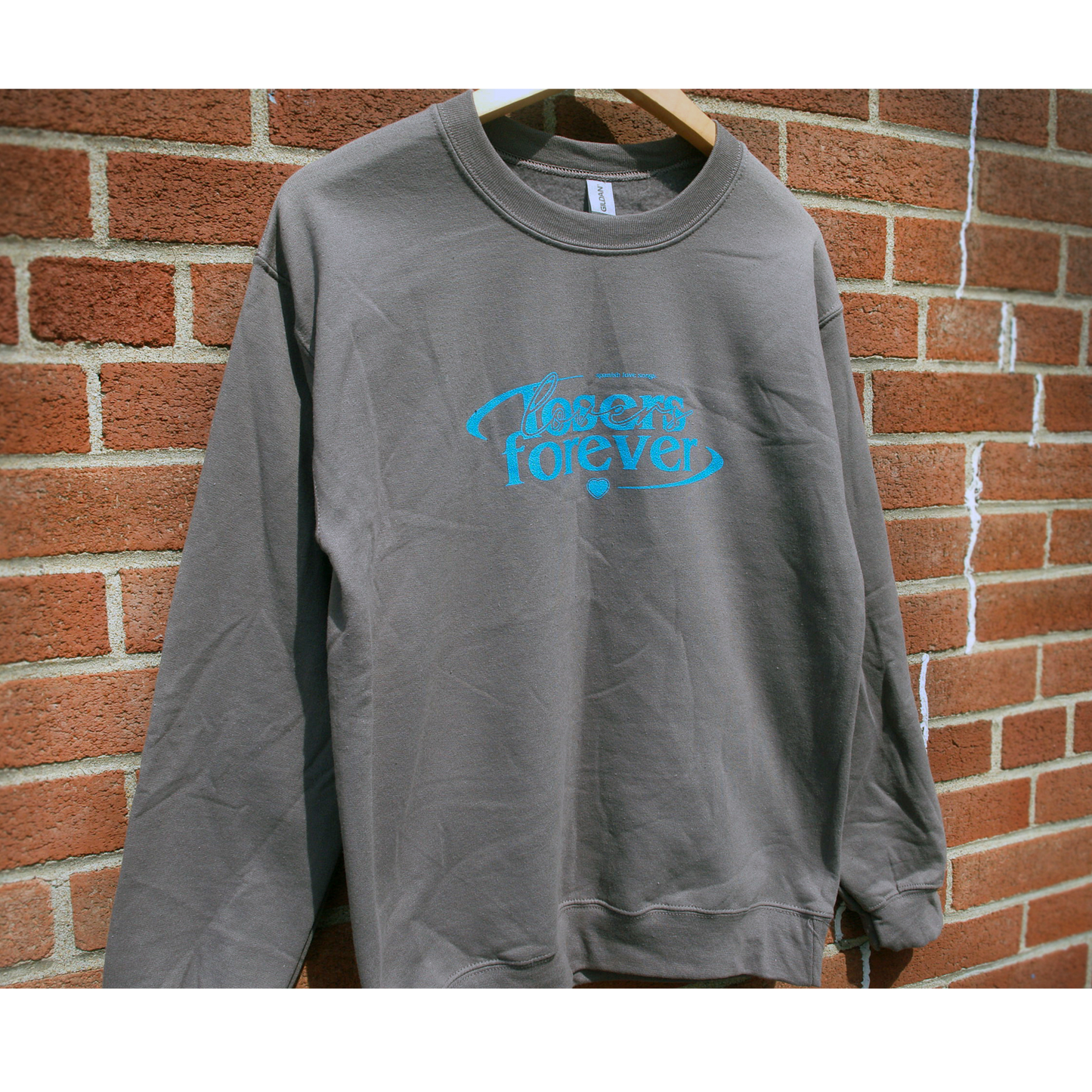 image of a charcoal crewneck sweatshirt hanging in front of a brick background. crewneck has center chest print in blue that says losers forever