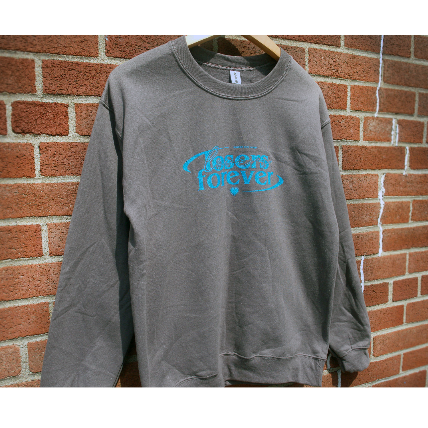 image of a charcoal crewneck sweatshirt hanging in front of a brick background. crewneck has center chest print in blue that says losers forever